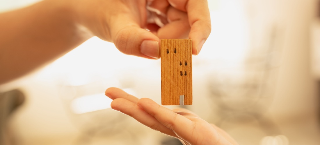 Una mano sostiene una casa de madera pequeña mientras otra la intenta coger por debajo